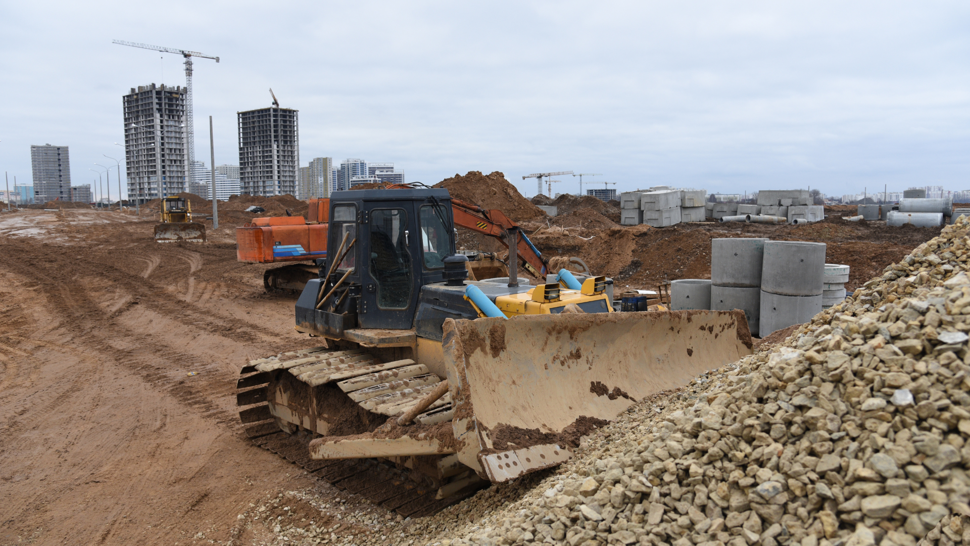 Bulldozer moving gravel at construction site; buildings under construction in the background.