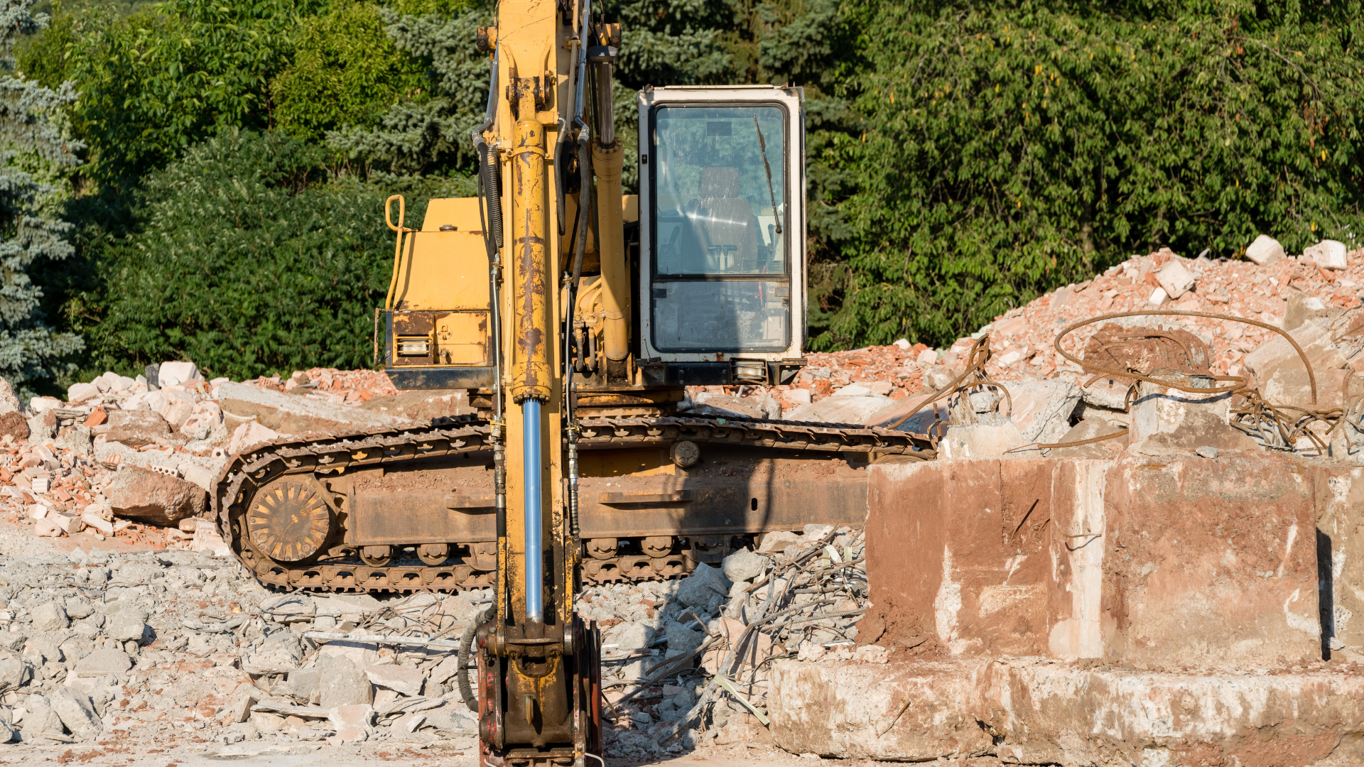 Yellow excavator demolishing a structure, surrounded by debris. Green trees in the background.