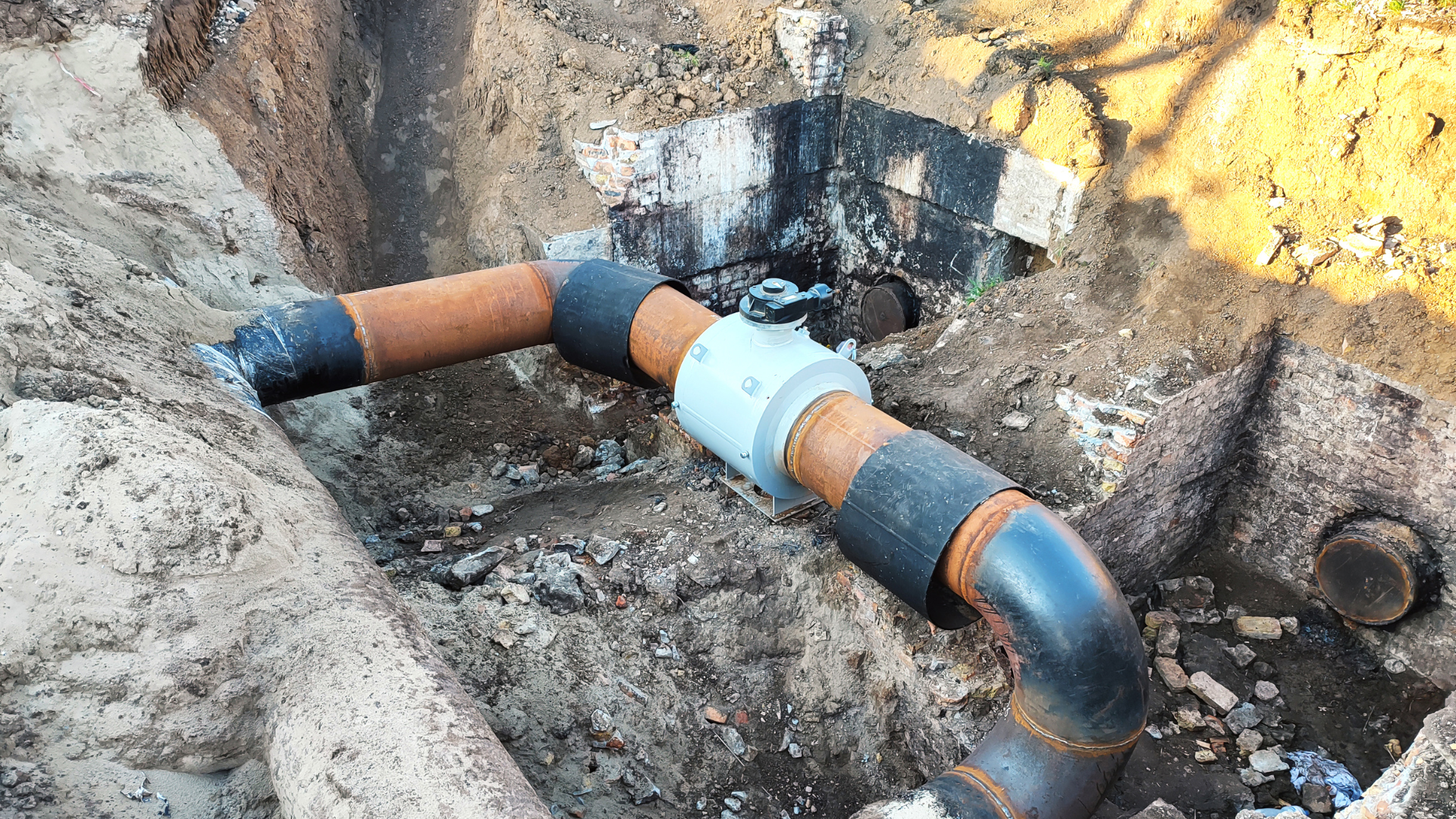 Black and brown pipes joined in a sand-filled trench.