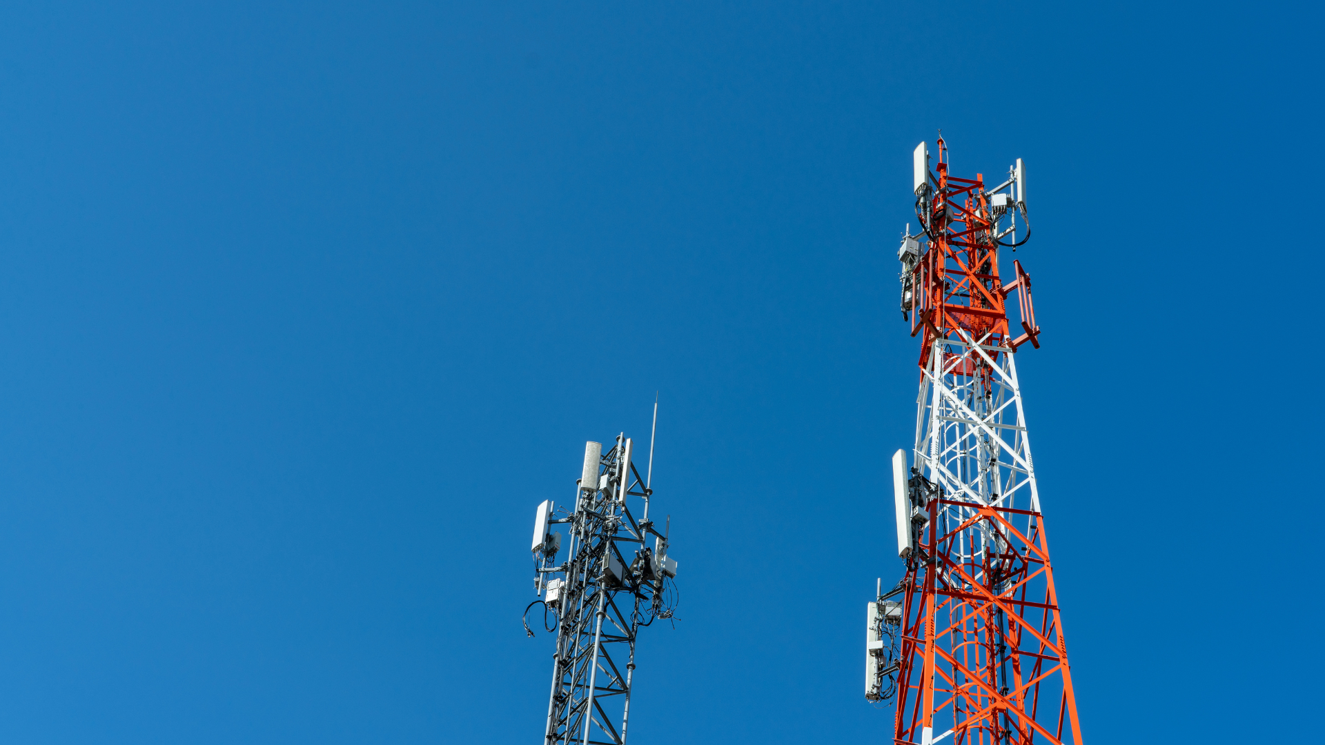 Two communication towers against a clear blue sky; one red and white, the other gray.