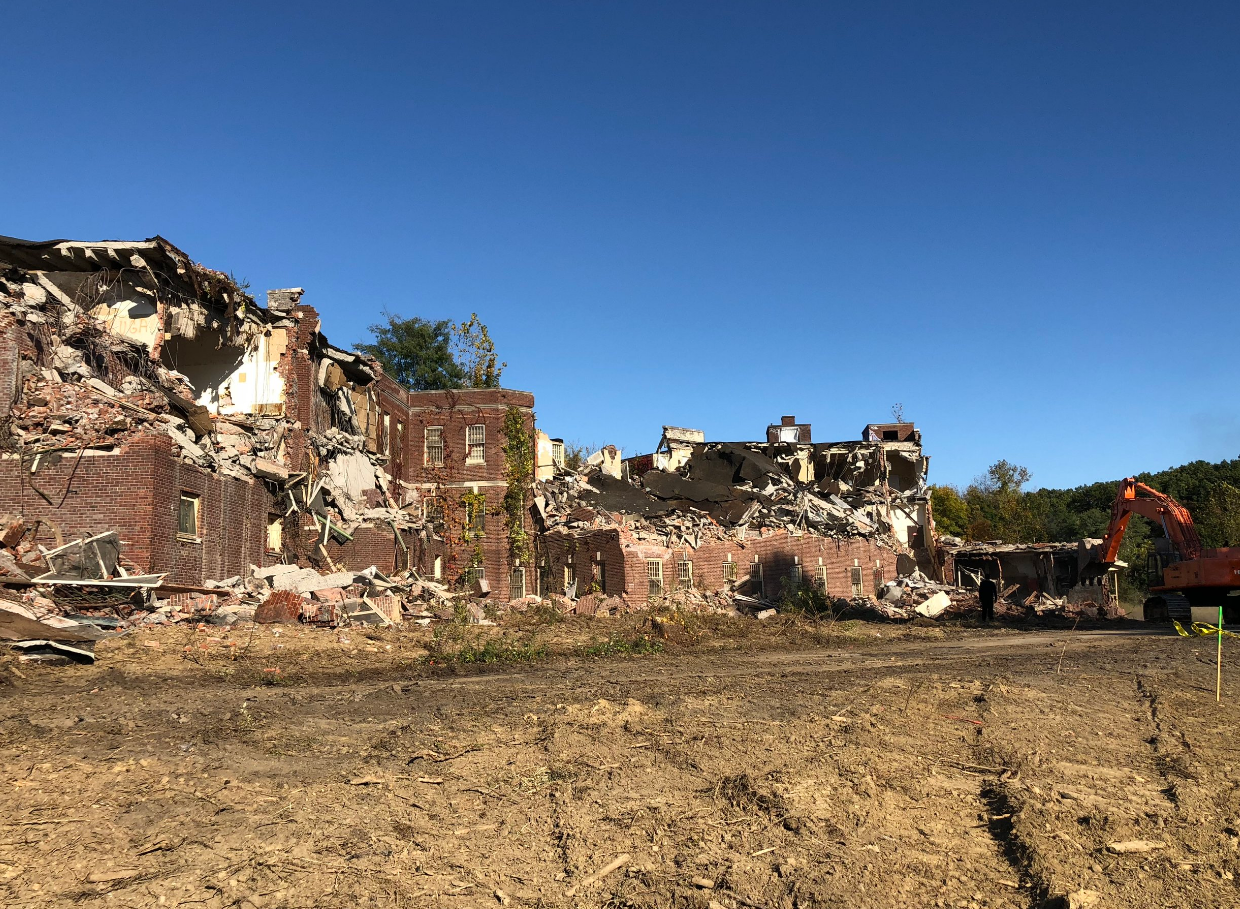 Demolished brick building under clear blue sky. Excavator visible, rubble on ground.