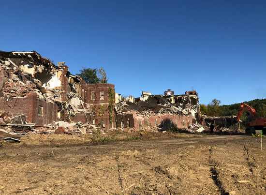 Demolished brick building under clear blue sky. Excavator visible, rubble on ground.