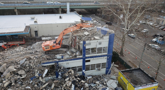 Demolition of a building by an orange excavator, debris piles surround the structure.