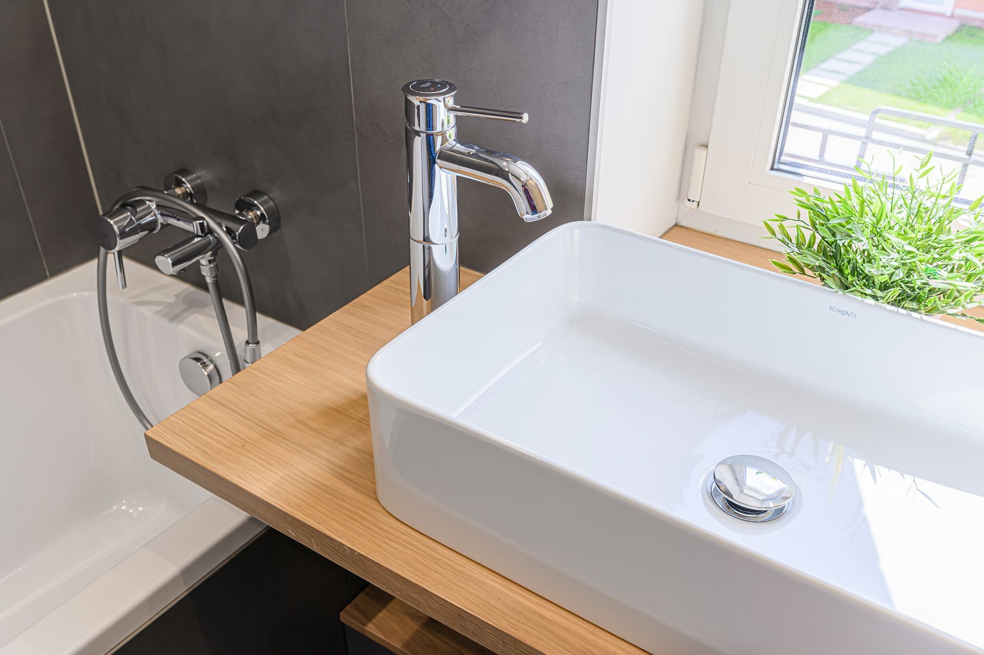 Modern bathroom sink with wooden countertop, chrome faucet, bathtub, and a window with green plants nearby