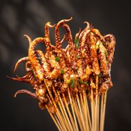 Grilled skewered octopus tentacles glazed with sauce and sprinkled with herbs, on a dark background
