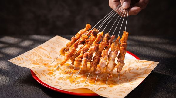 Hand holding skewers over a sauce-coated square plate on a dark table
