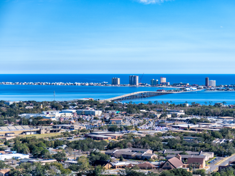 Coastal cityscape with homes, a bridge, blue ocean, and distant high-rise buildings under a clear sky