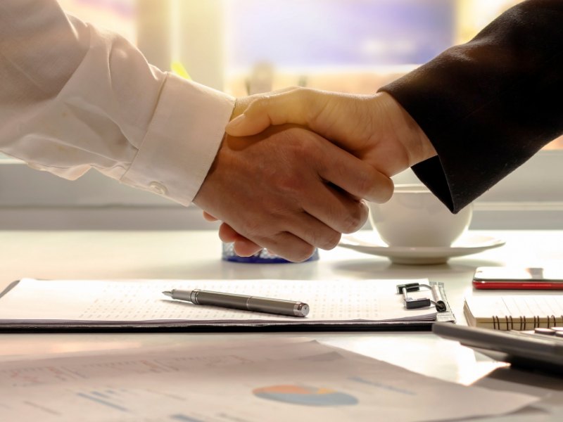 Two people shake hands across a desk with documents and a pen in a bright office.