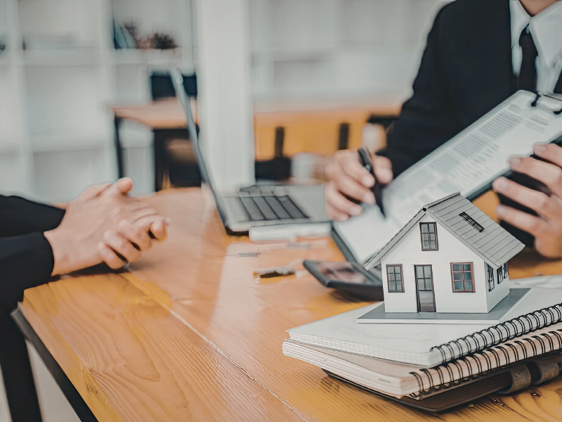Business meeting with hands around a laptop, paperwork, and a small model house on a wooden table