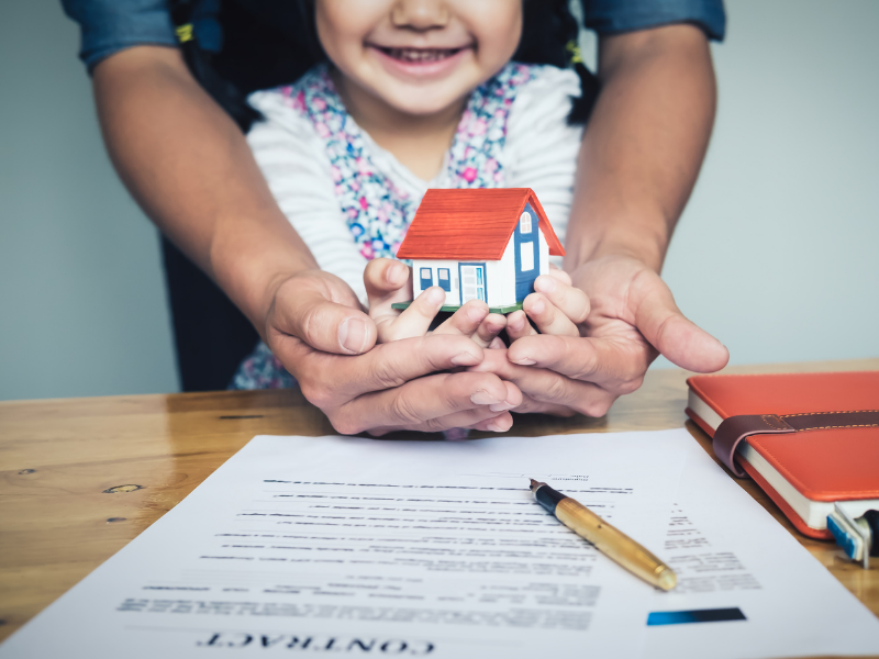 Child smiling behind hands holding a small toy house over a desk with paperwork and a pen