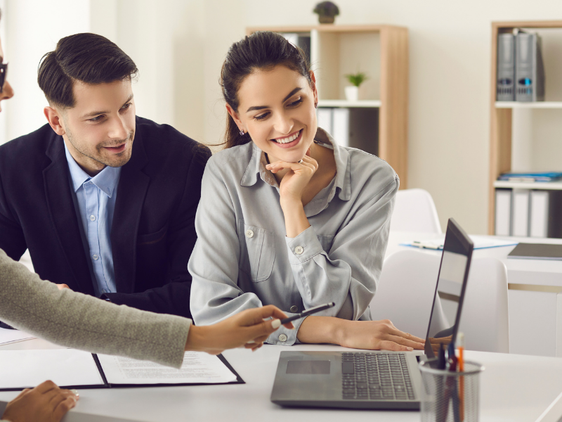 Two coworkers reviewing documents at a desk in a bright office, smiling and discussing a laptop screen