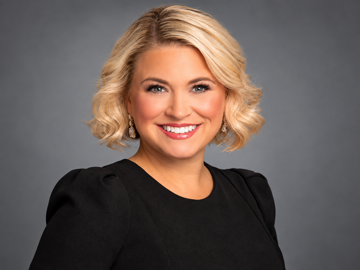Smiling person with short blonde hair wearing a black top against a gray background