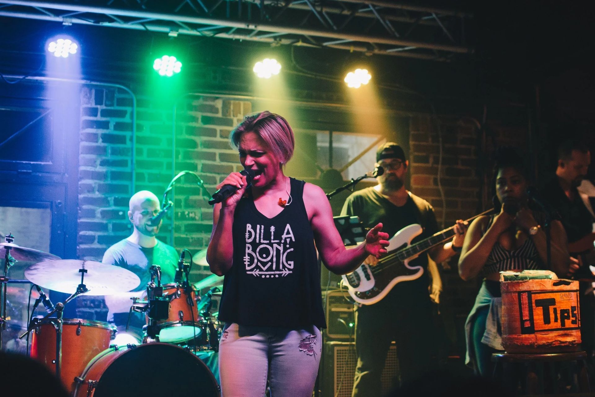 A band performing on stage; singer with microphone, drummer, bassist, and horn player. Colorful stage lighting.