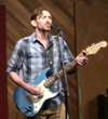 Man playing a blue electric guitar and singing into a microphone on stage, wearing a plaid shirt.