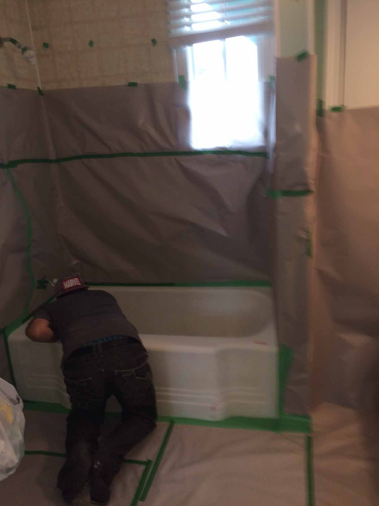 Une personne penchée au-dessus d'une baignoire blanche dans une salle de bains en rénovation, recouverte de bâches en plastique et de ruban adhésif.