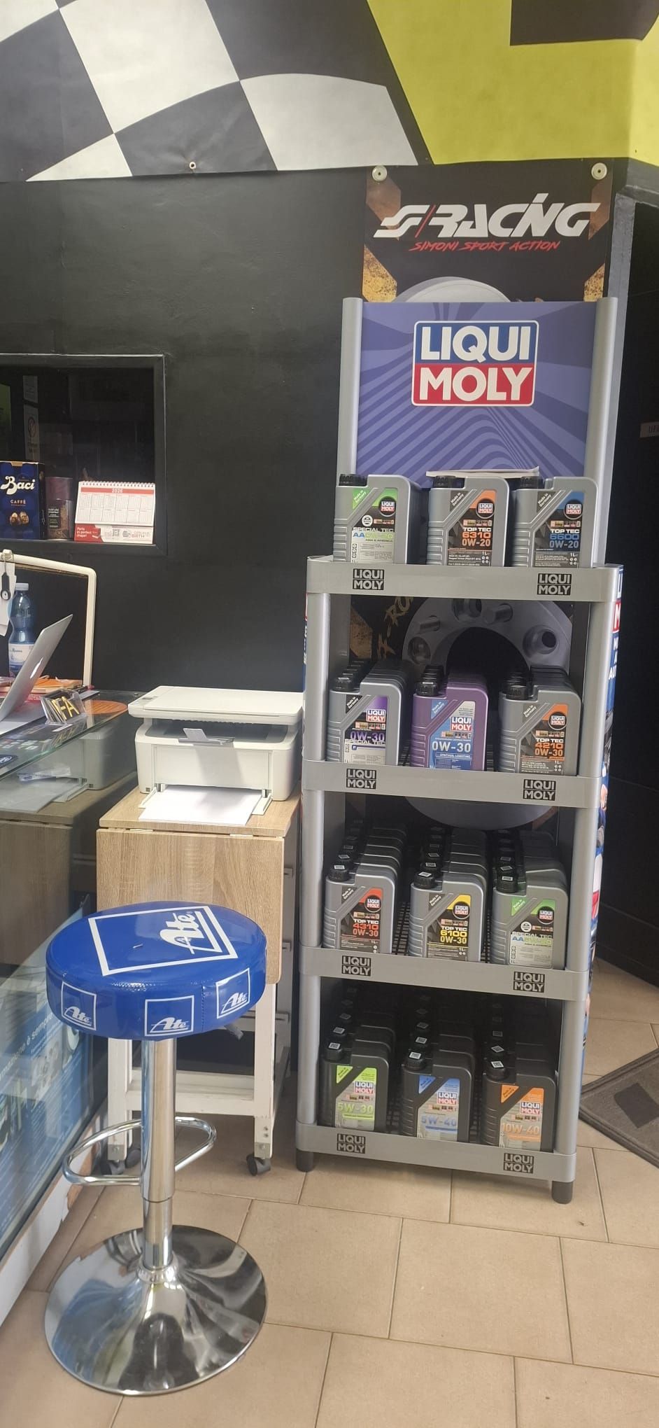 Esposizione di prodotti Liqui Moly accanto a uno sgabello blu in un interno di negozio piastrellato.