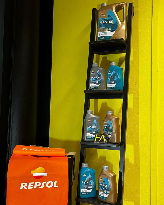 Scaffale di bottiglie di olio motore accanto a un contenitore arancione della Repsol, contro una parete gialla.