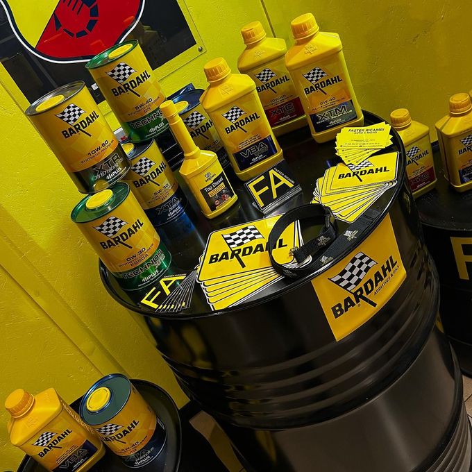 Contenitori gialli per olio Bardahl impilati su barili neri in un luminoso stand espositivo.