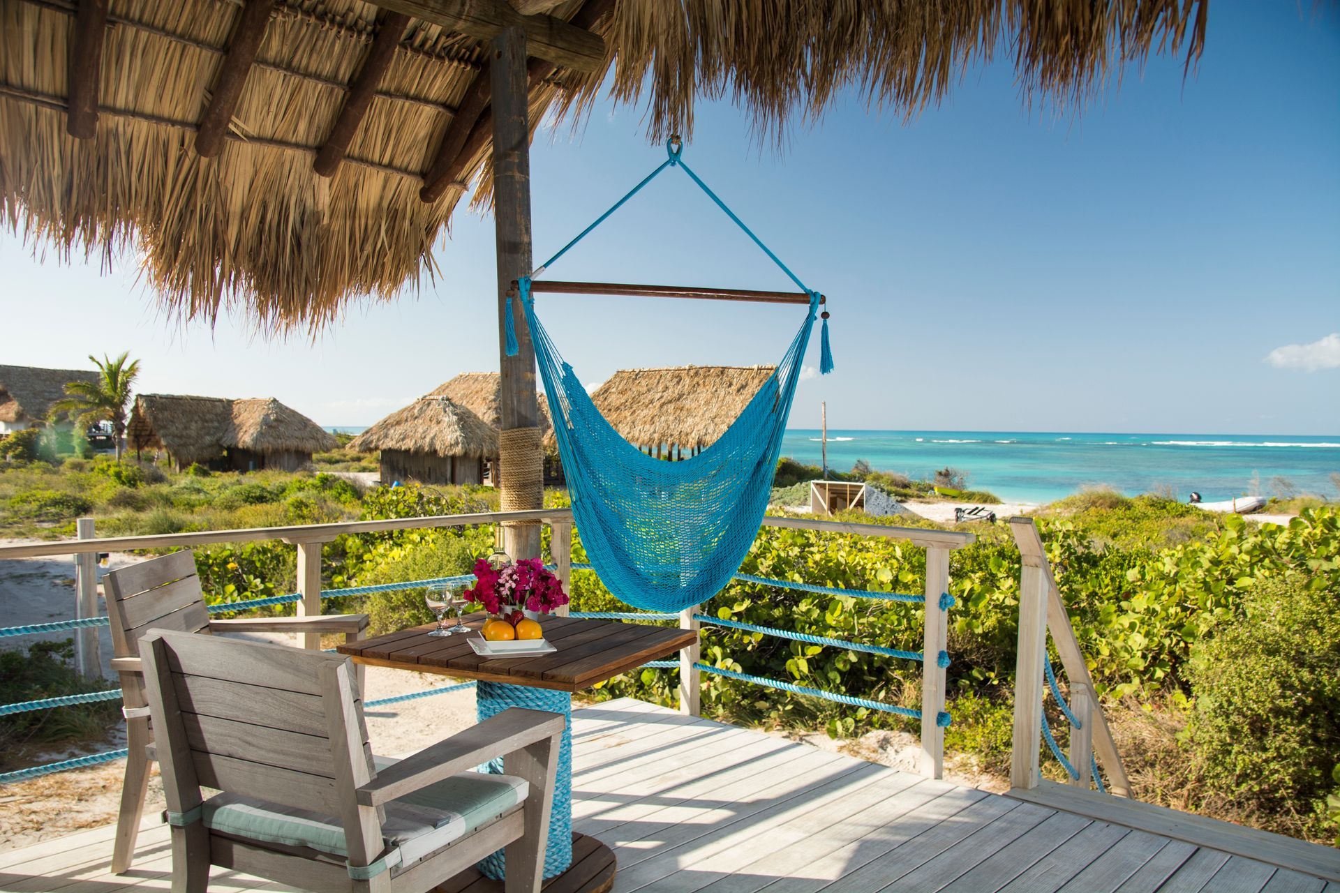 Wooden patio with a blue hammock, table with flowers, thatched roof, and ocean view.