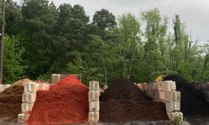 Three large piles of red, brown, and black mulch stacked beside a wooded area
