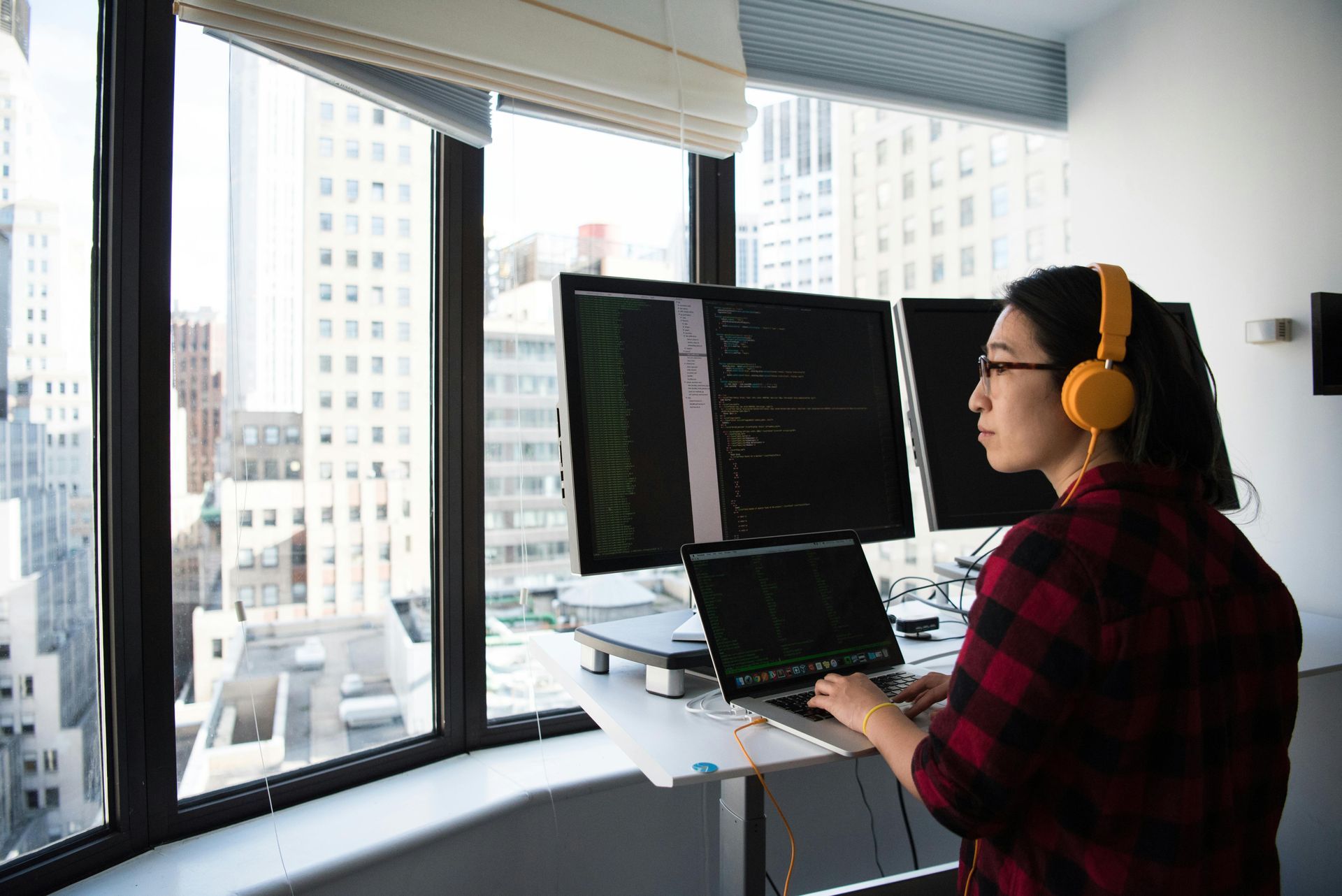 Woman in headphones coding at standing desk near a window overlooking a city.
