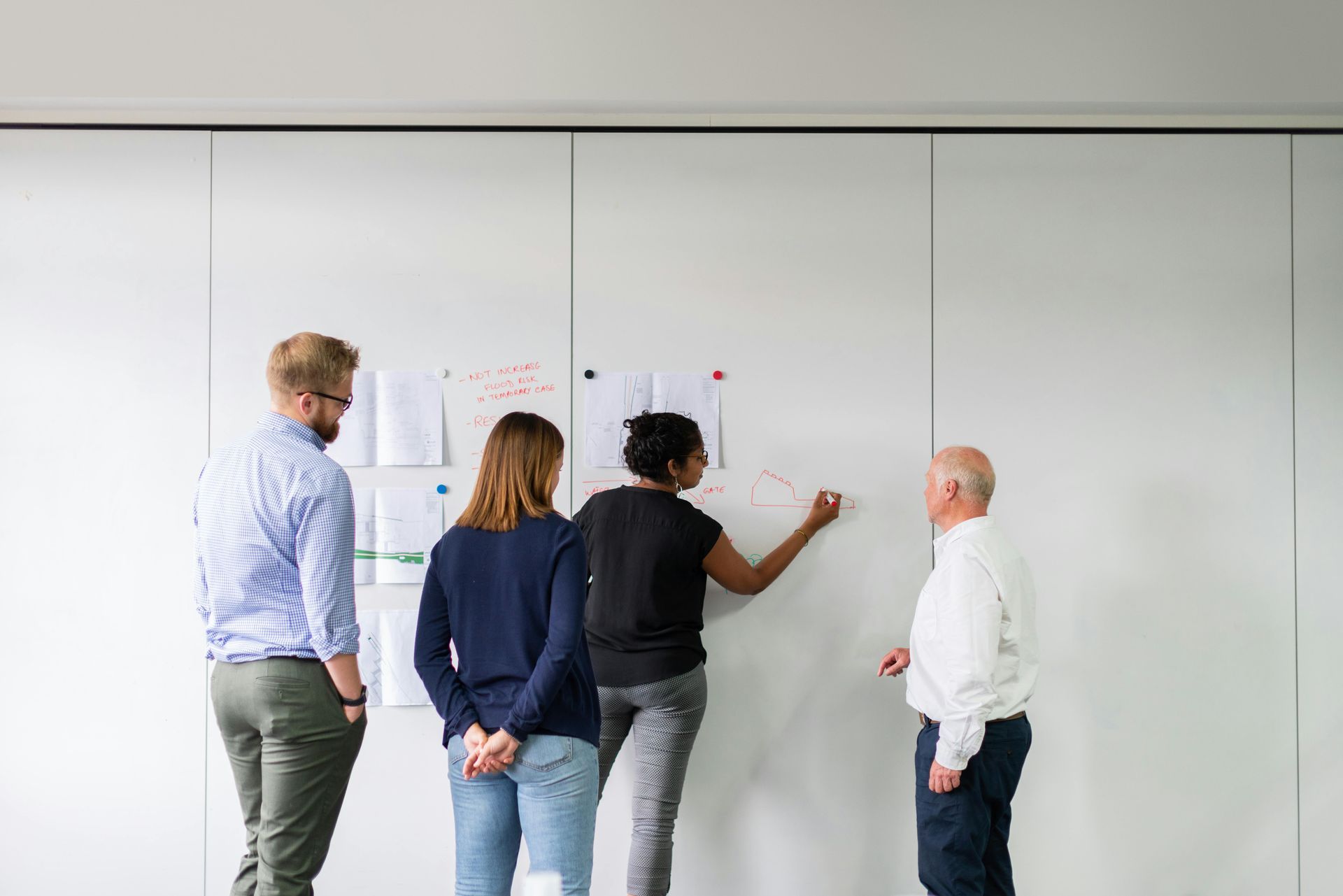Four people collaborating, looking at a whiteboard with notes and diagrams, one writing with a marker.