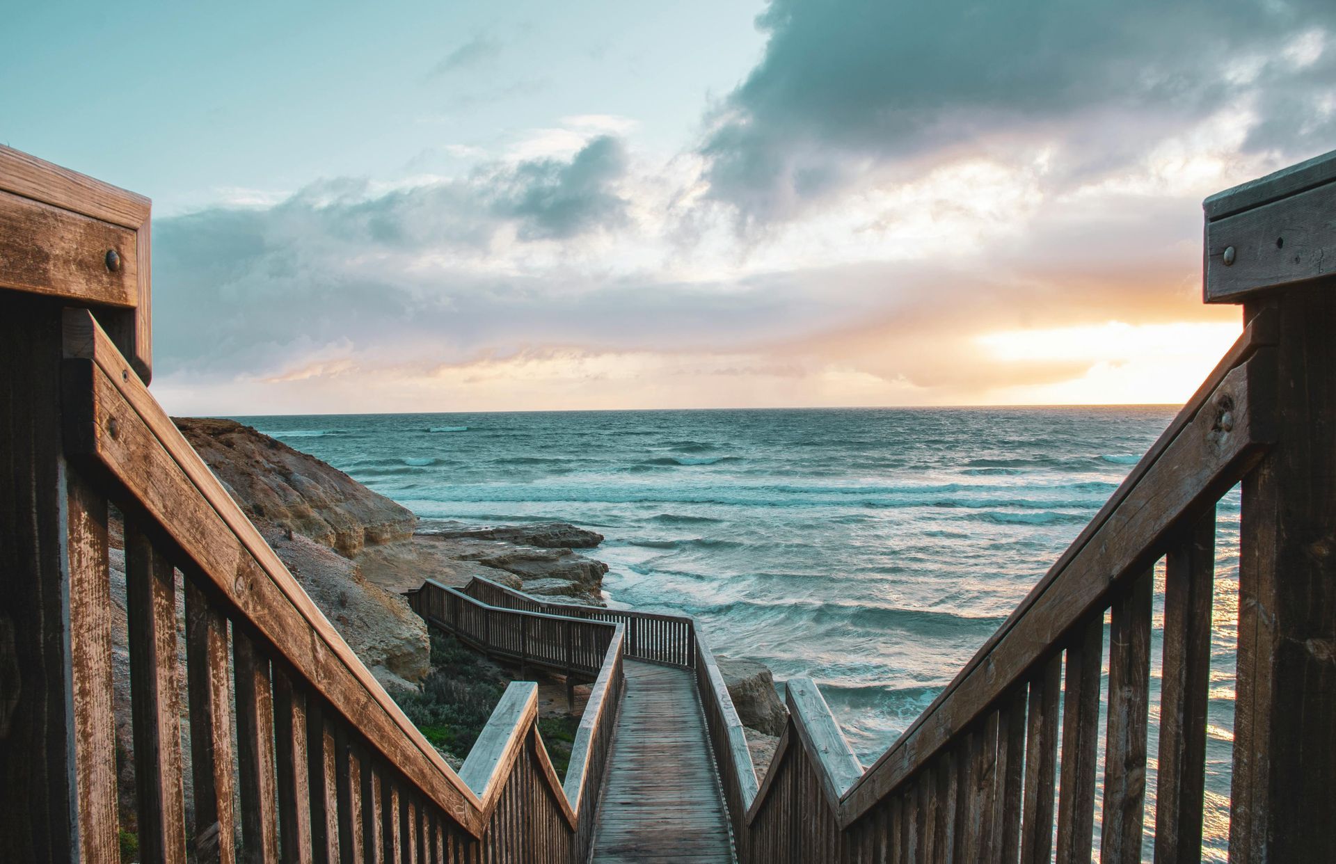 Wooden stairs leading to the ocean at sunset, with cloudy sky and waves crashing on the shore.