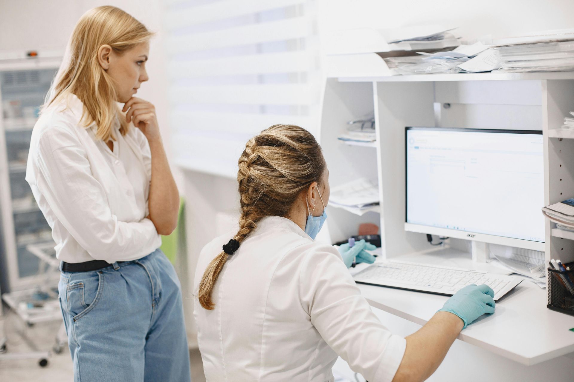 Woman in jeans watches as a healthcare worker in a mask and gloves types on a computer.