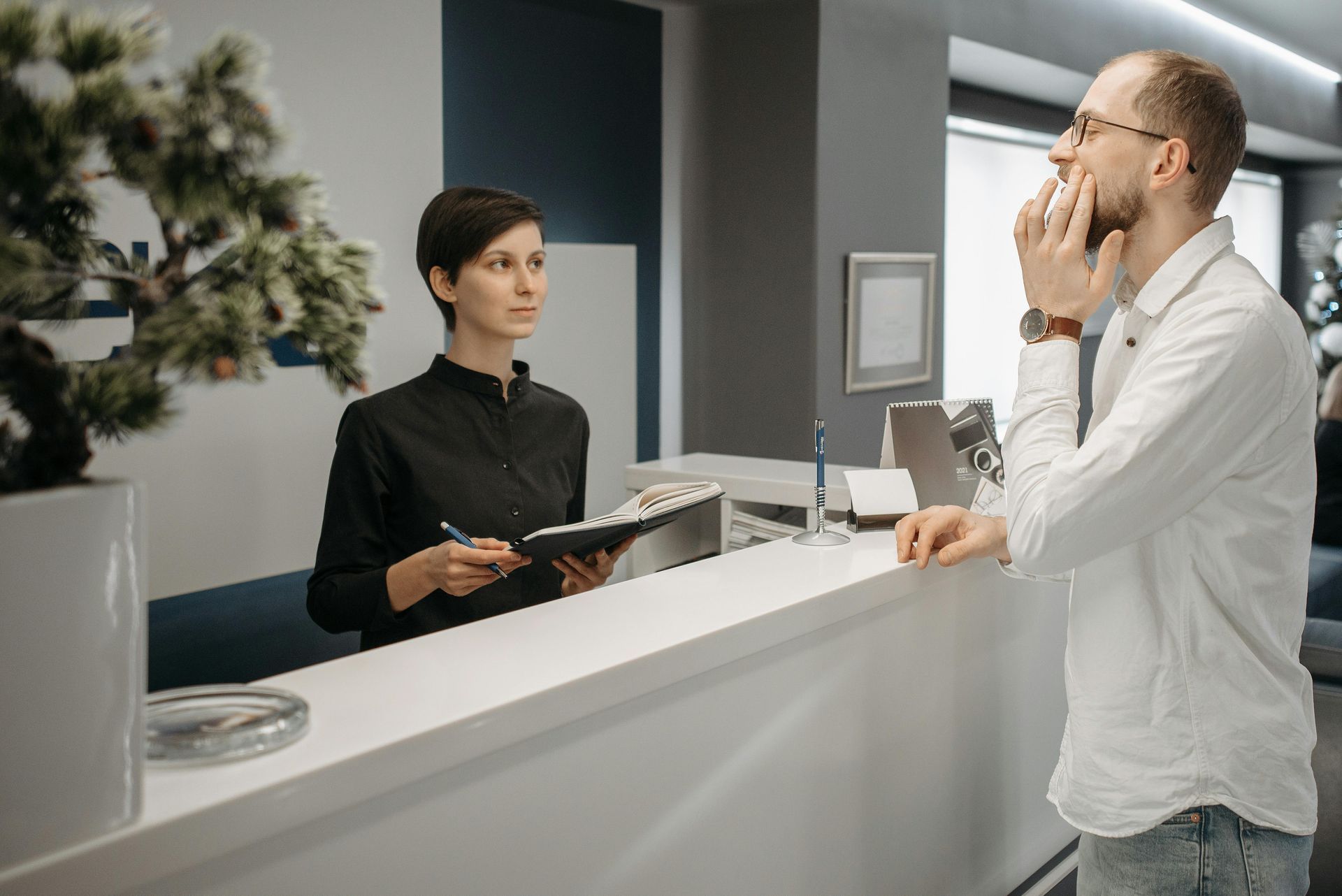 Woman at desk, writing, talking to a man with glasses, touching his face, in an office reception area.