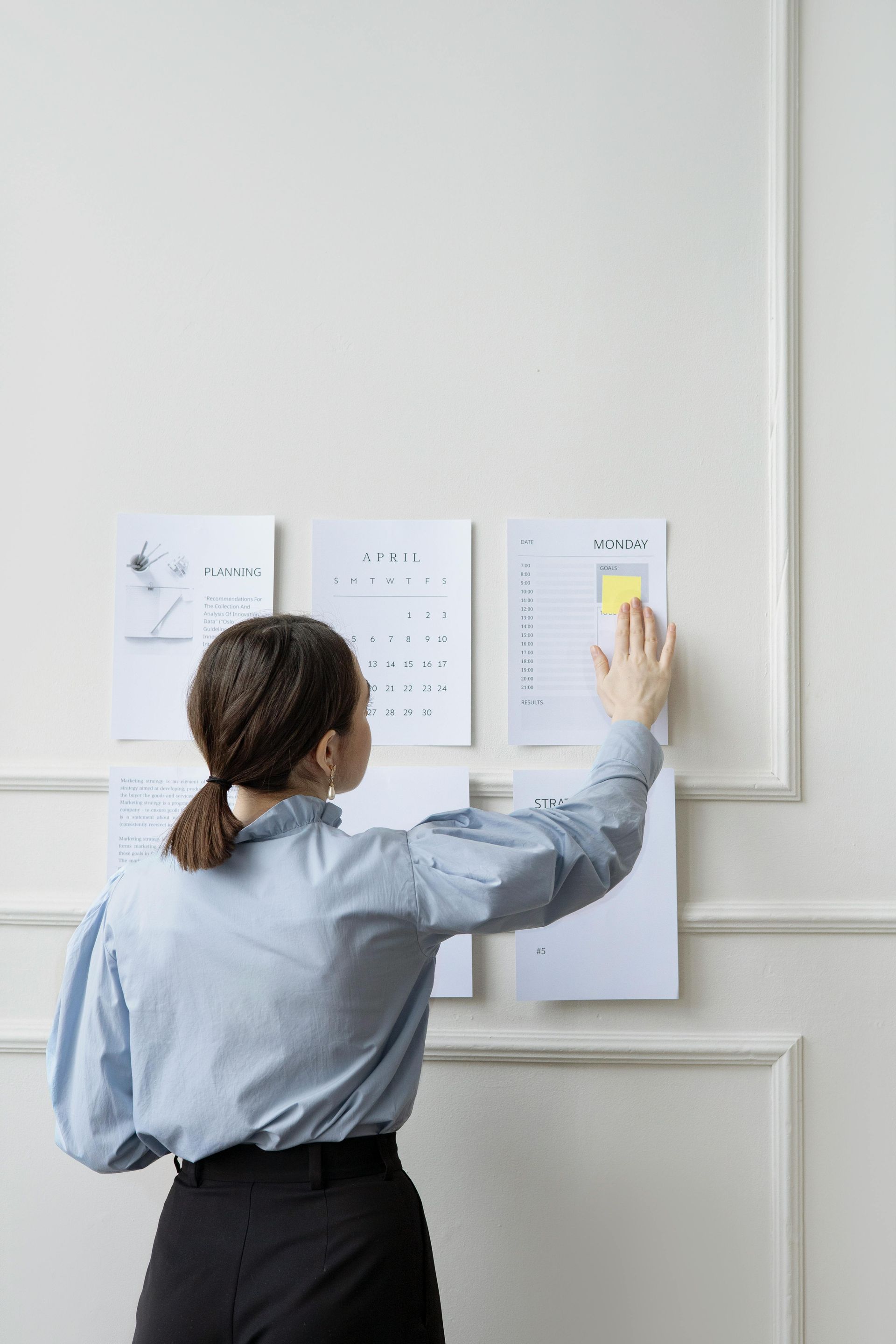 Woman in blue shirt, black pants, posting yellow sticky note on papers mounted on white wall.