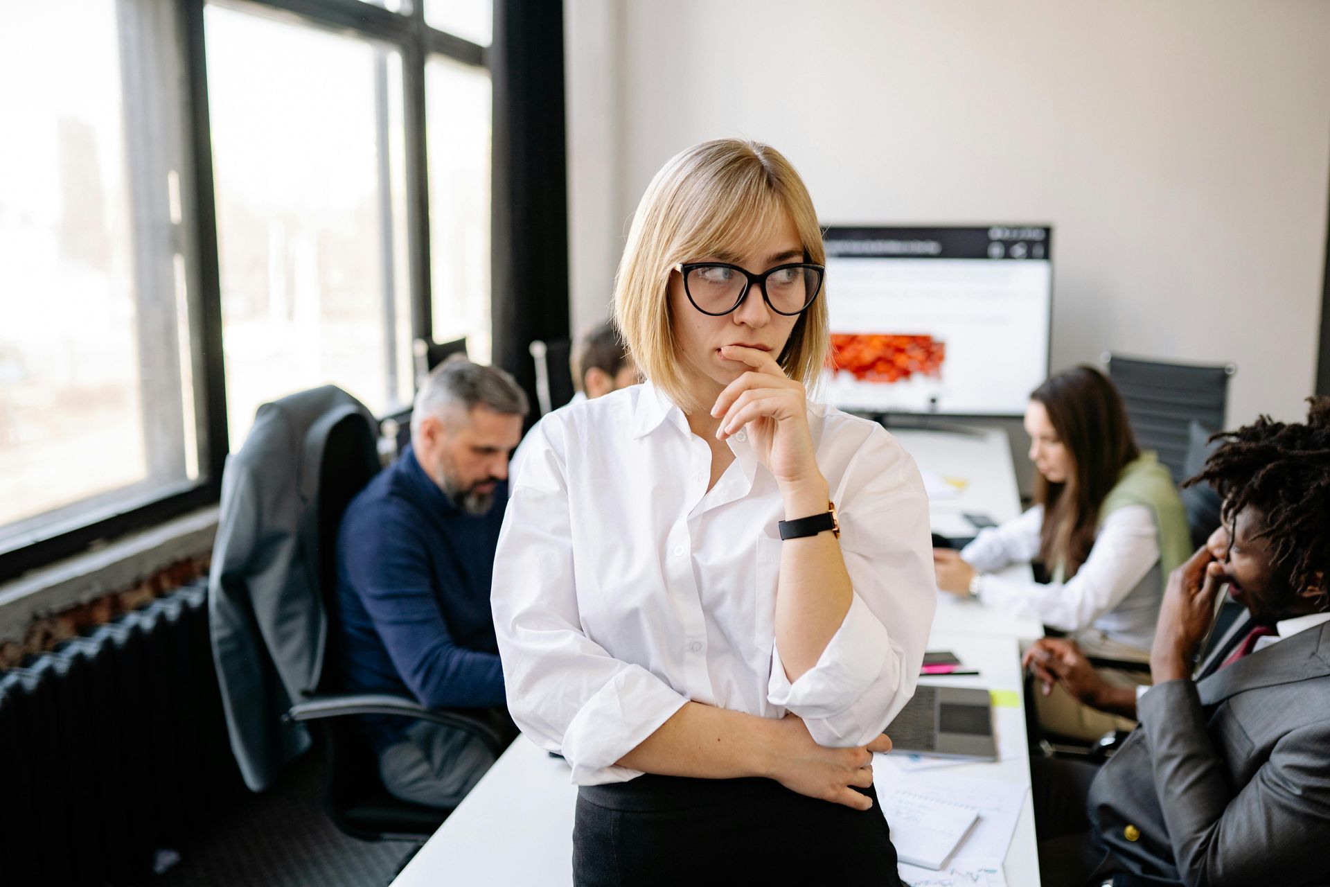 Woman in glasses pondering, in a modern office with colleagues in the background.