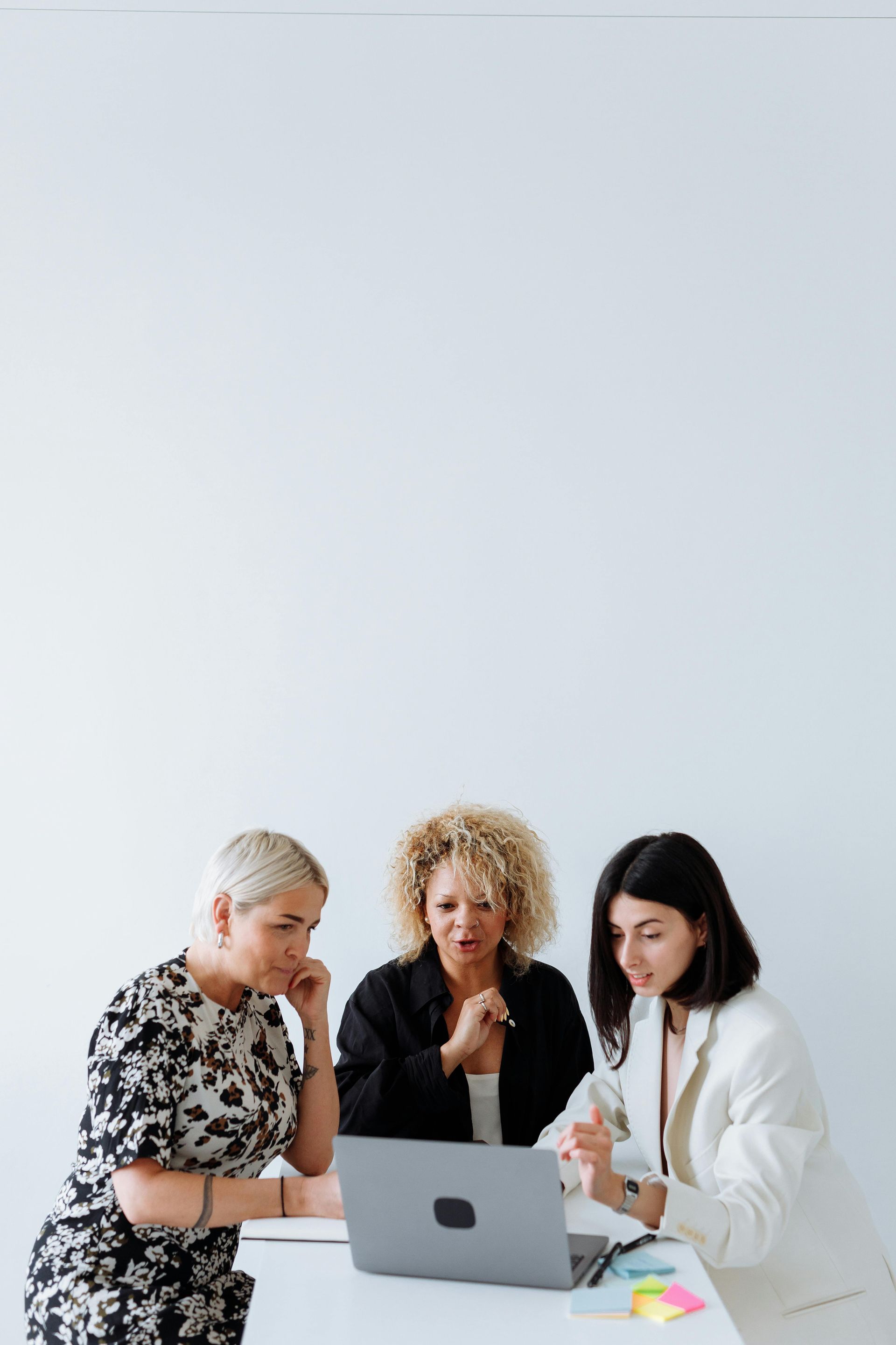 Three women at a table, looking at a laptop. One points, the others lean in with expressions of interest. White background.