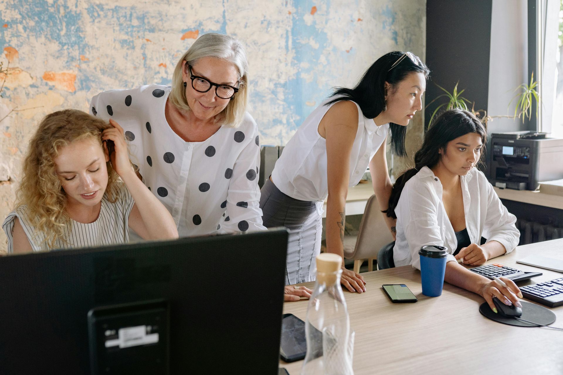 Four women collaborating around a computer in an office. One is using a mouse.