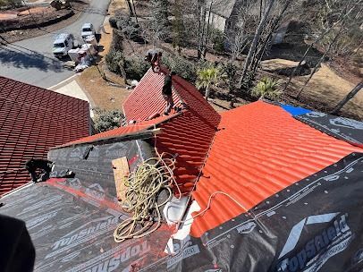 Roofers working on a red metal roof, debris, and tools visible. Sunny day with vehicles in the background.