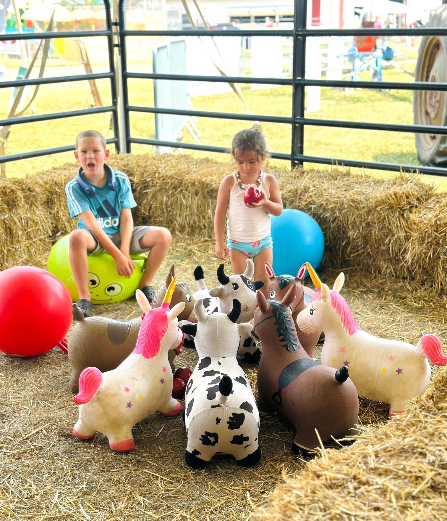 Children playing with inflatable hopper toys, including unicorns and a cow, in a hay-filled pen.