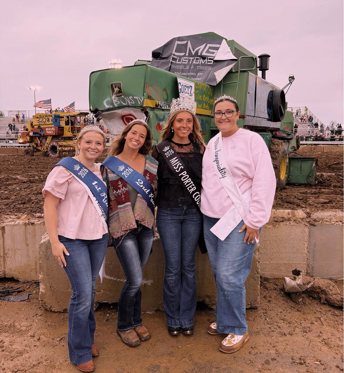 Four women smiling, wearing crowns and sashes, holding a guitar, a shoe, and a hat. Black and white photo.