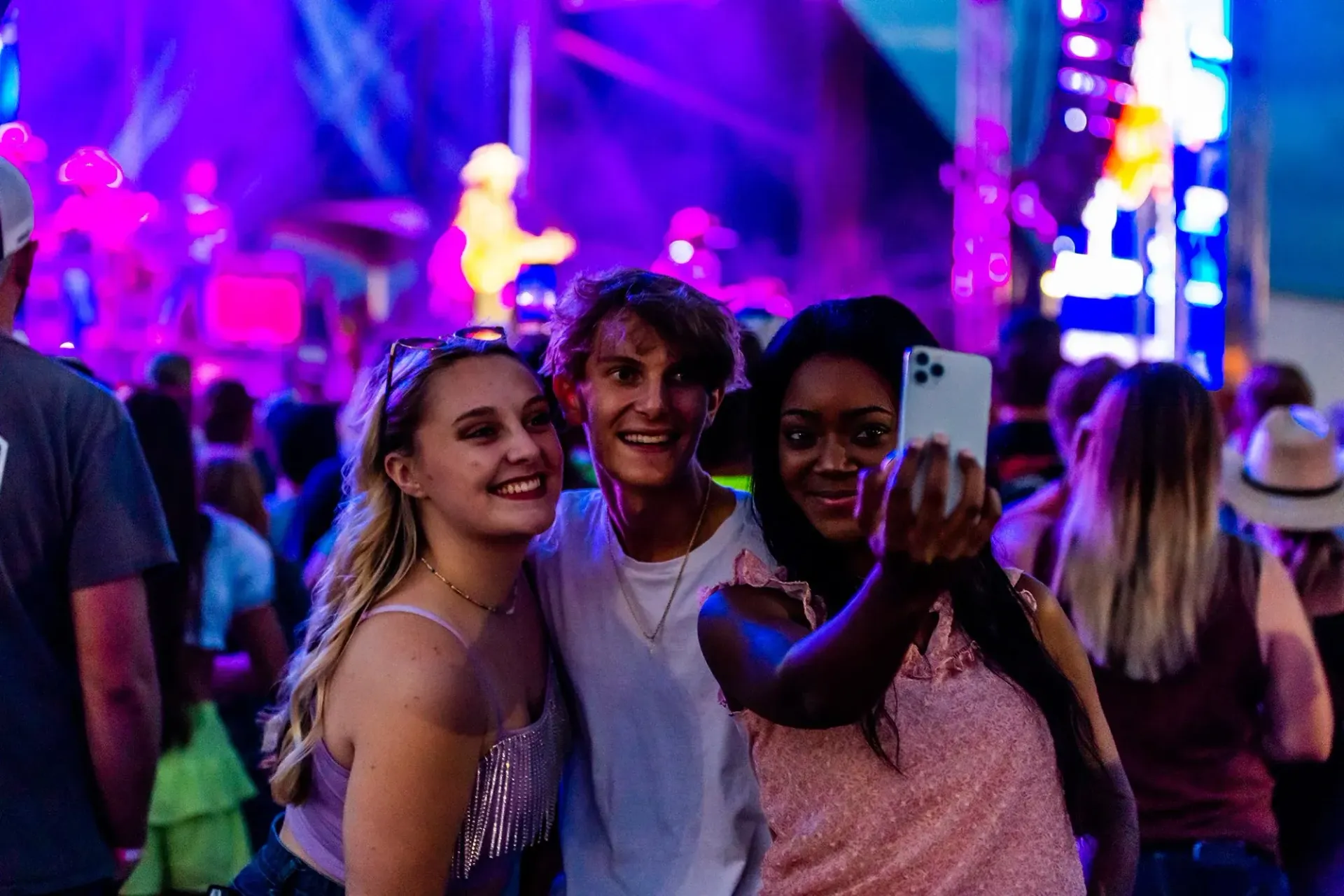 Three people take a selfie at a brightly lit outdoor music event.
