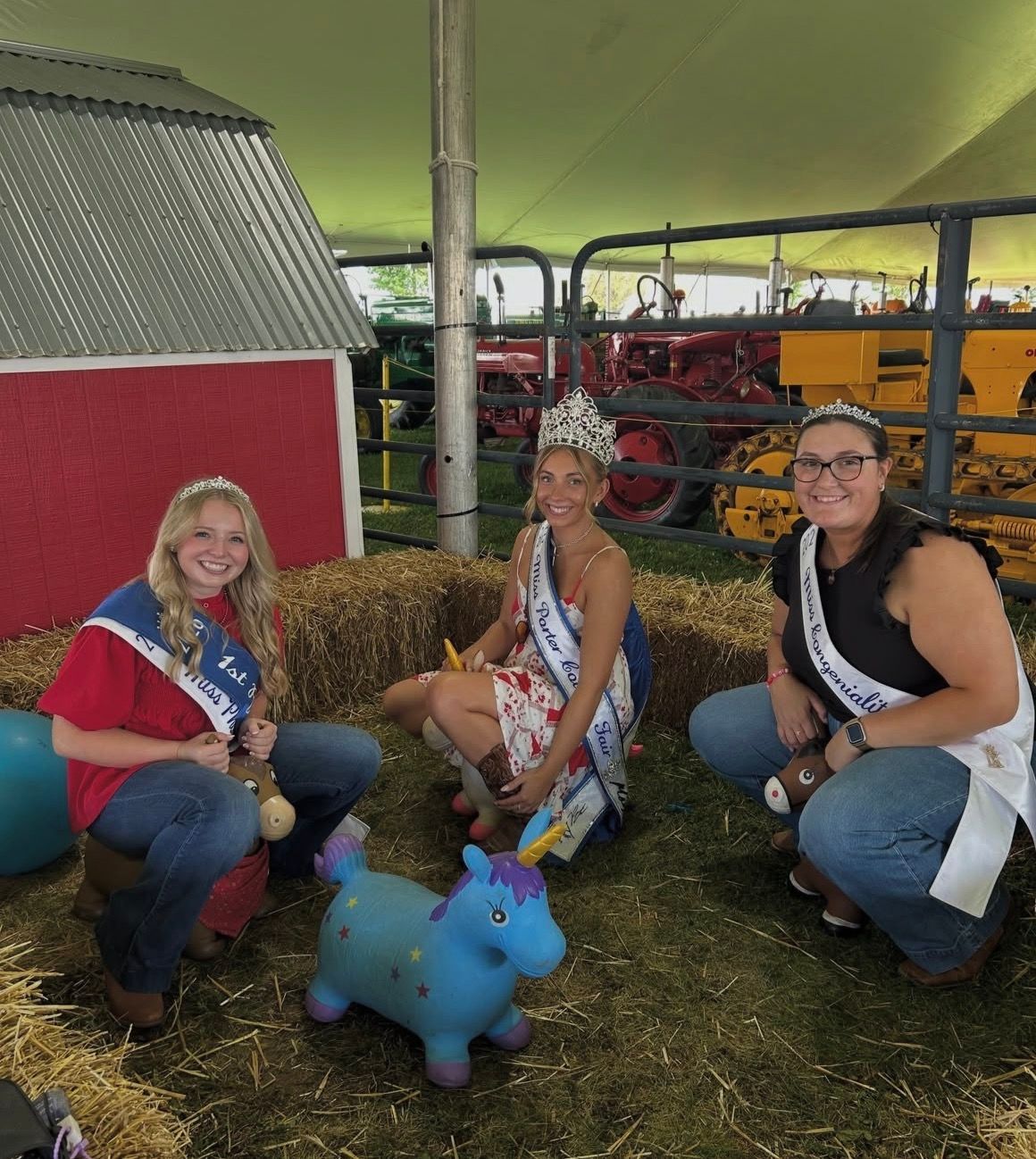 Four women in crowns and sashes pose with a tennis shoe, guitar, and trophy at a stage.