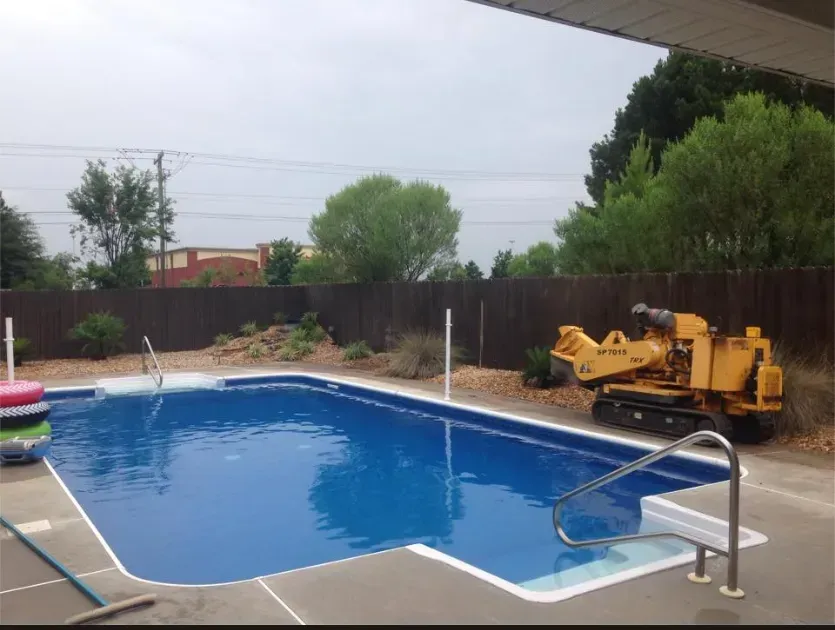 A backyard pool with blue water. A stump grinder sits on the edge. A brown fence surrounds the yard.