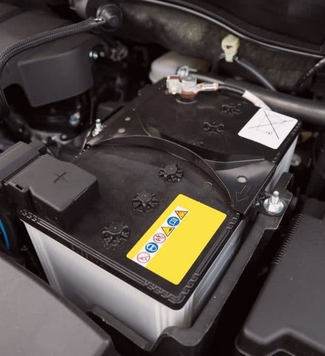 Car battery in engine compartment with warning label.