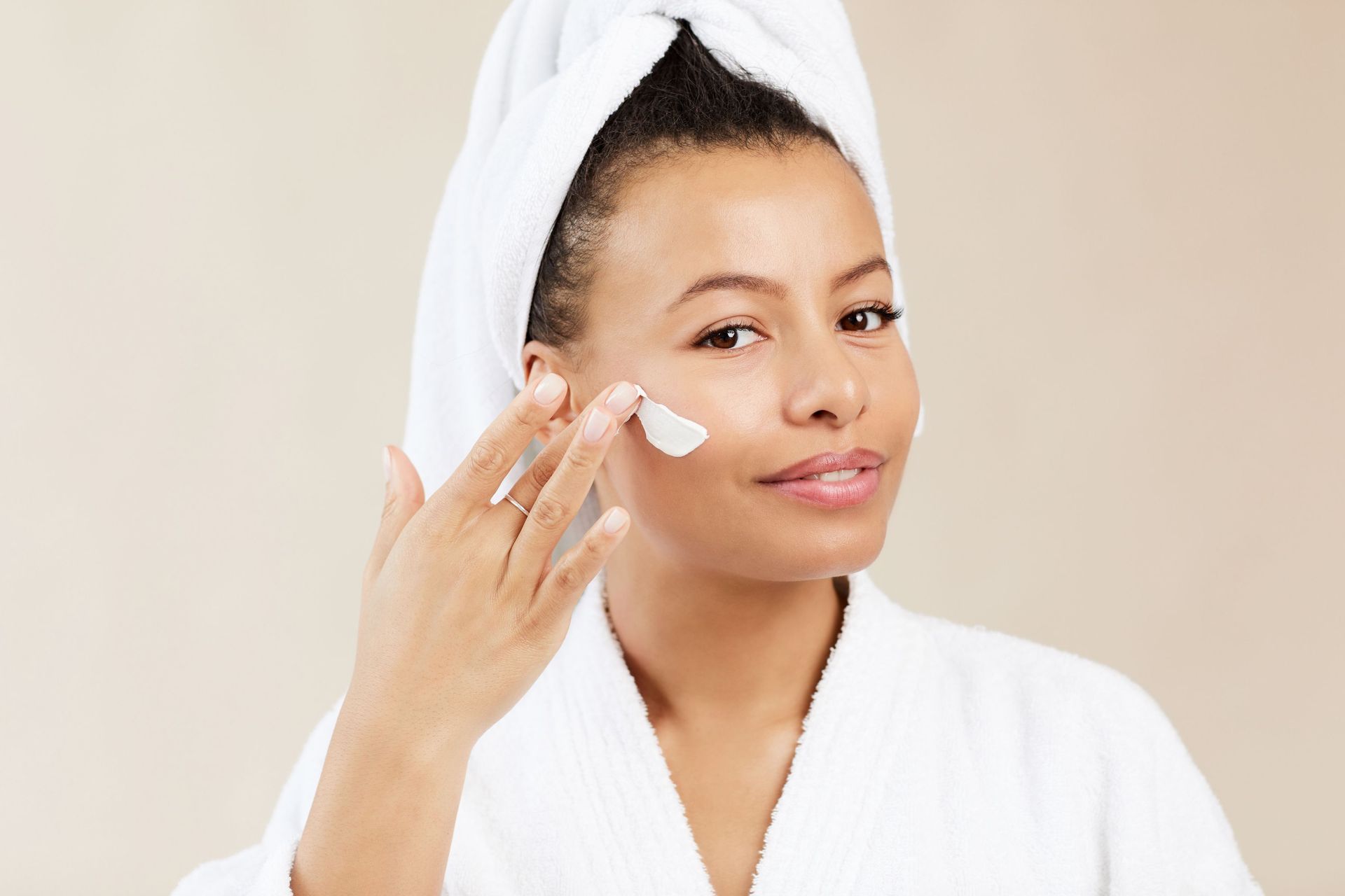 A woman with a towel wrapped around her head is applying lotion to her face.