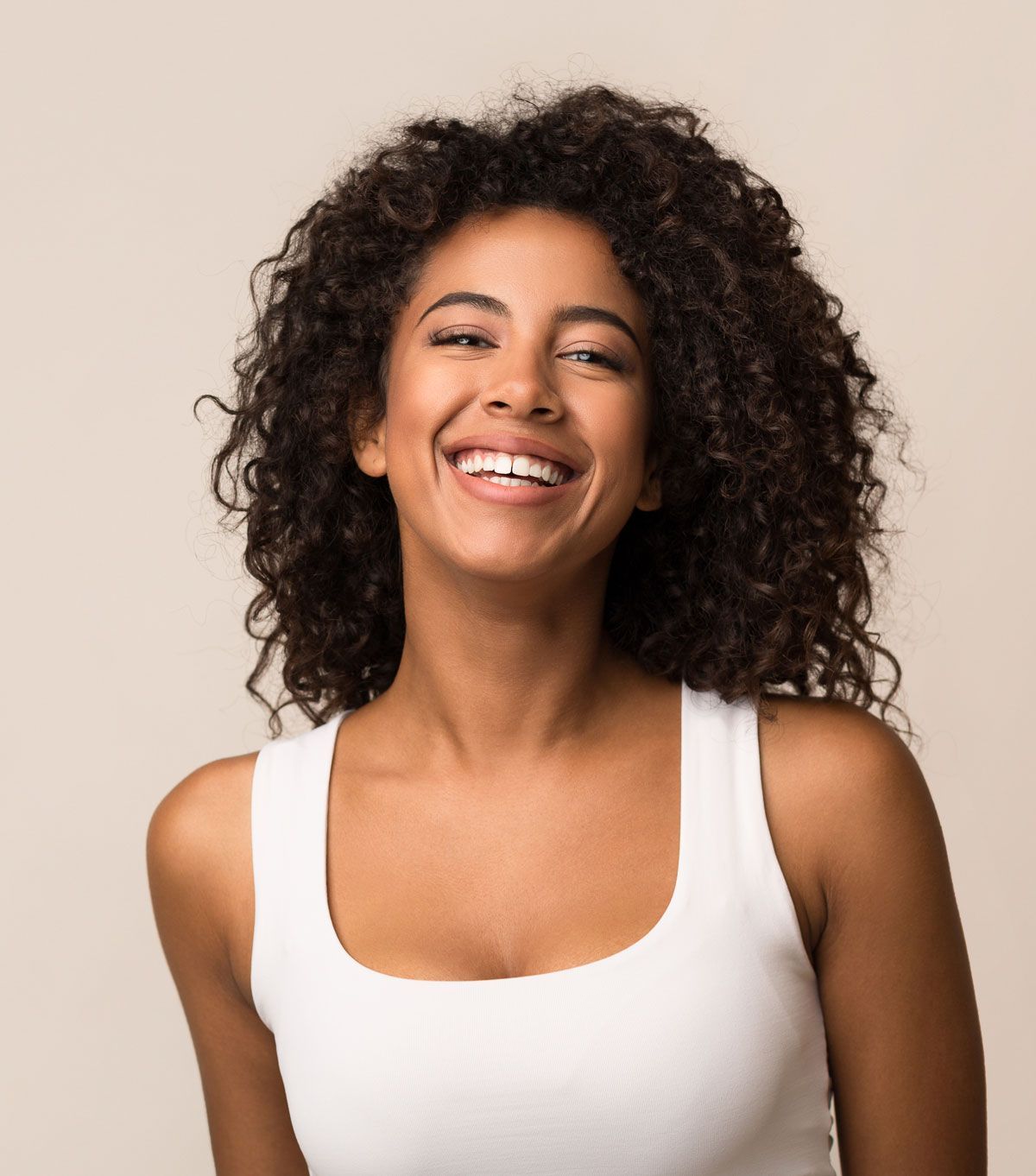 A woman with curly hair is smiling and wearing a white tank top.