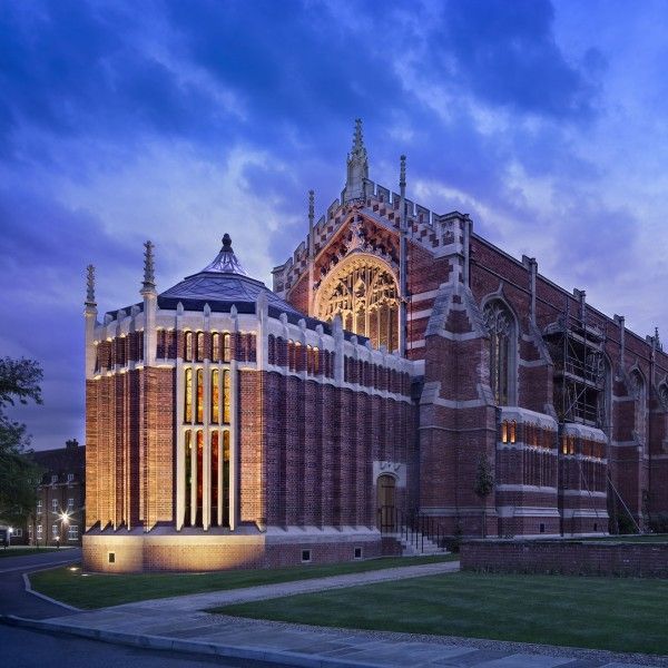 Gothic red-brick chapel with glowing arched entrance at dusk under a blue sky.