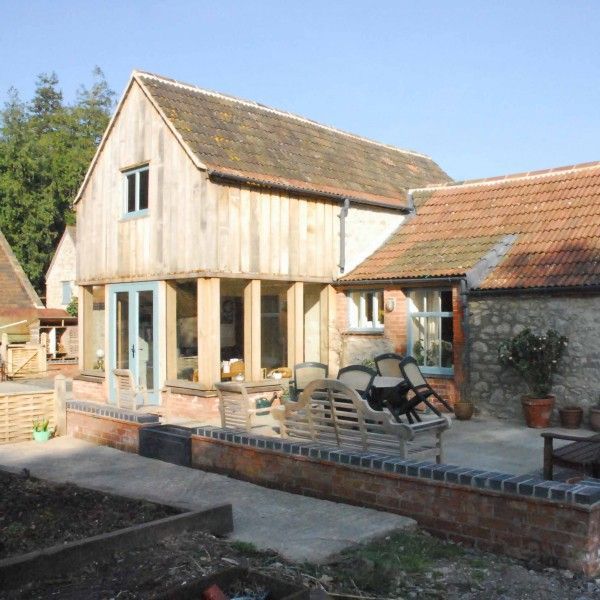 Rustic stone and wood house with large windows, patio seating, and tiled roof in a sunny courtyard