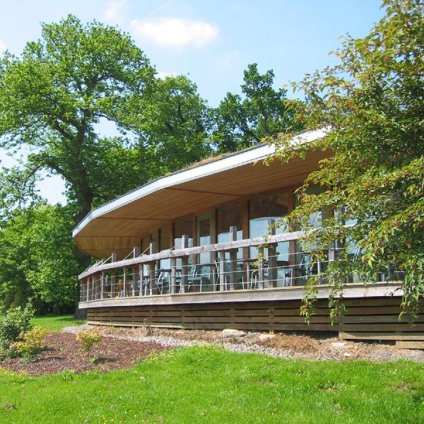 Modern wooden building with curved roof and glass railing, surrounded by green trees and lawn