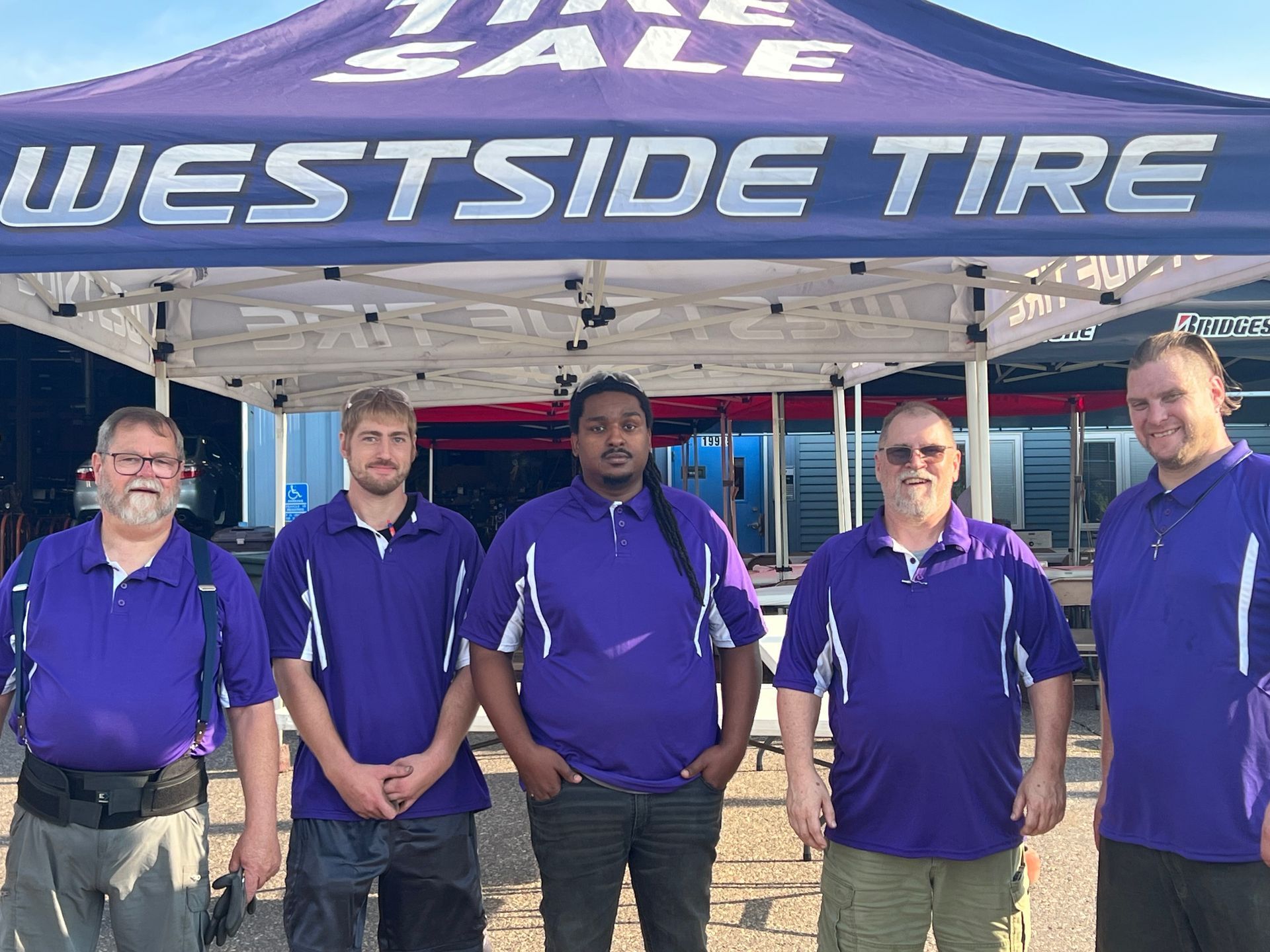 Five men in purple shirts standing under a Westside Tire canopy outdoors