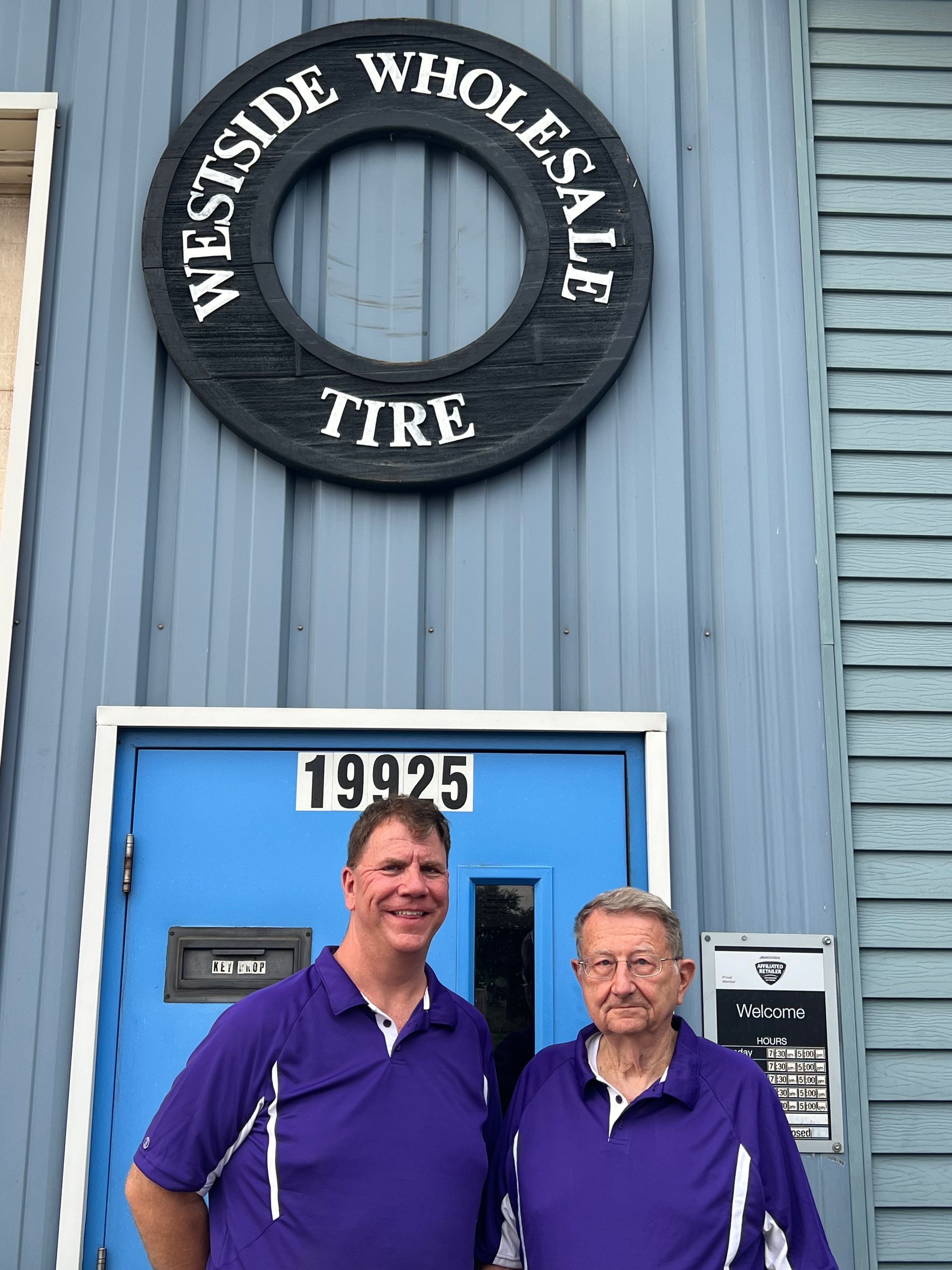 Two people stand outside WesSide Wholesale Tire beneath a blue door and circular shop sign.