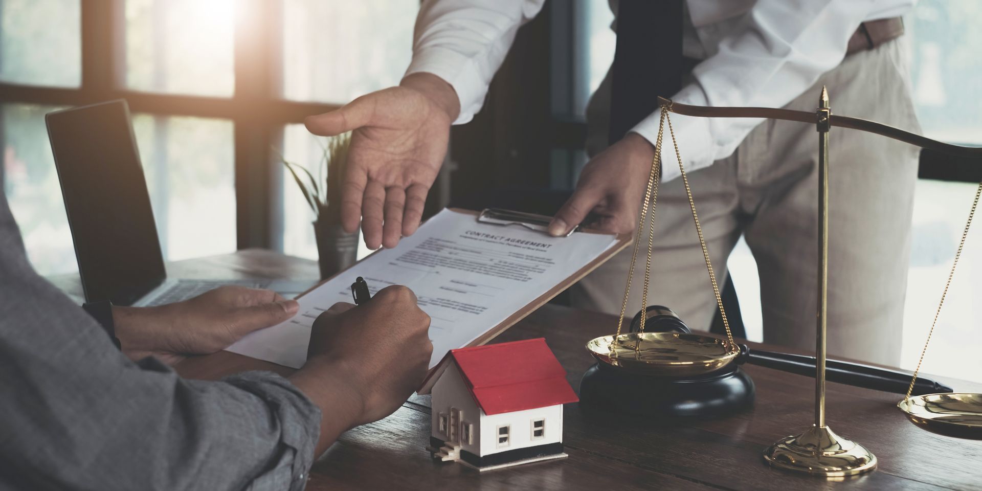 Client signing legal documents with a bankruptcy attorney, scales, and a house model on the desk.