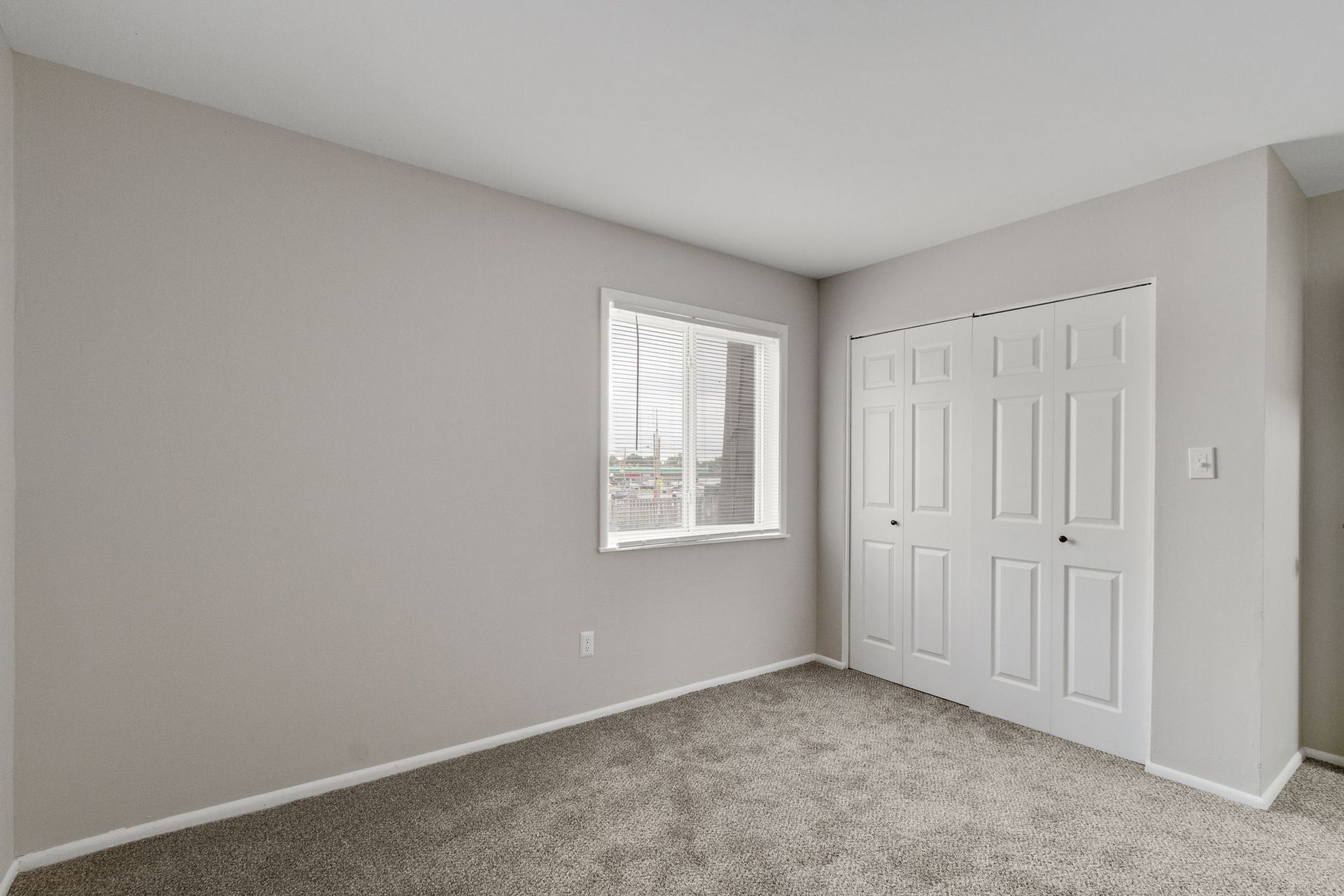 Empty bedroom with gray walls, carpet, window, and white closet doors.
