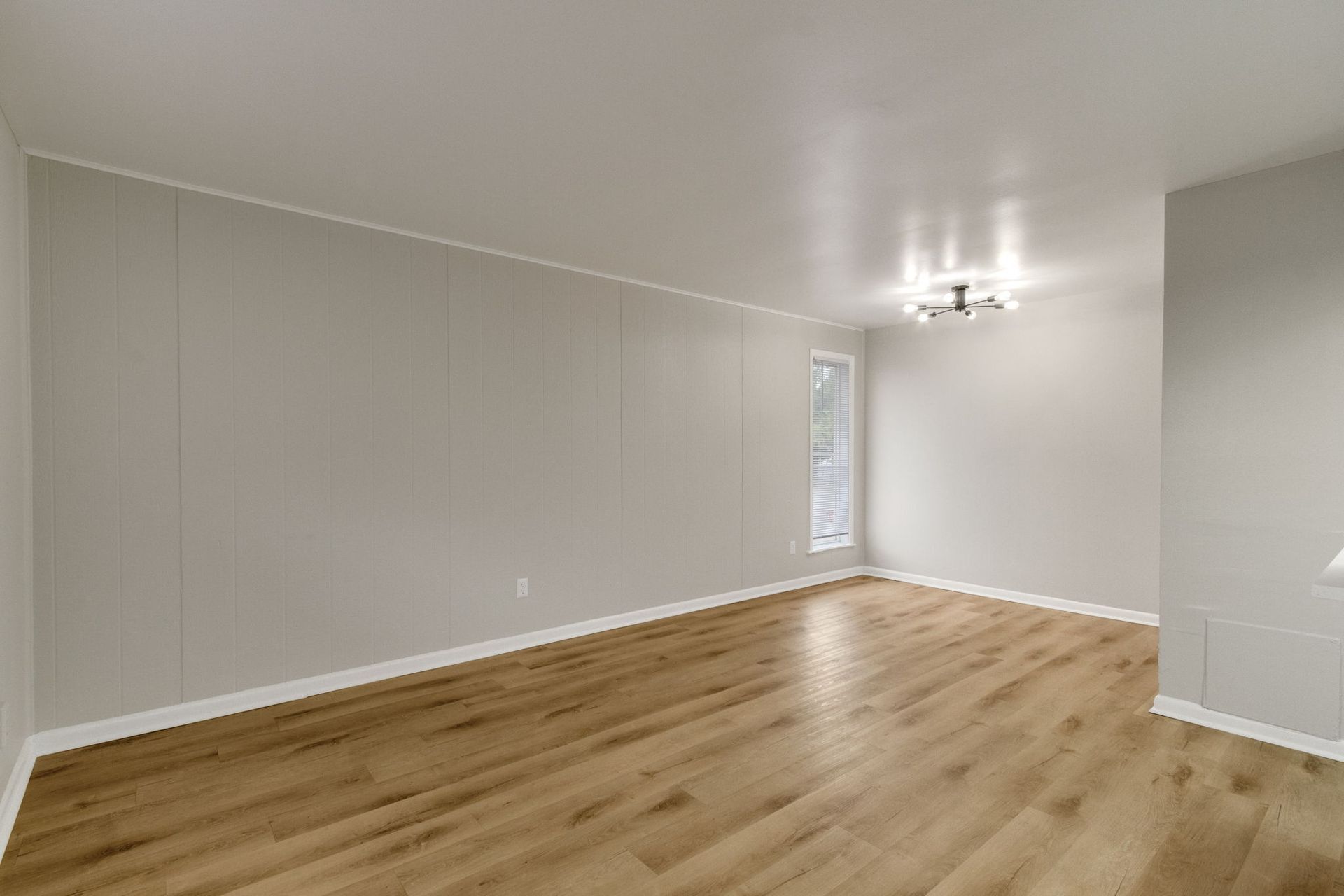 Empty room with light wooden floors and grey walls, a small window, and a ceiling light fixture.
